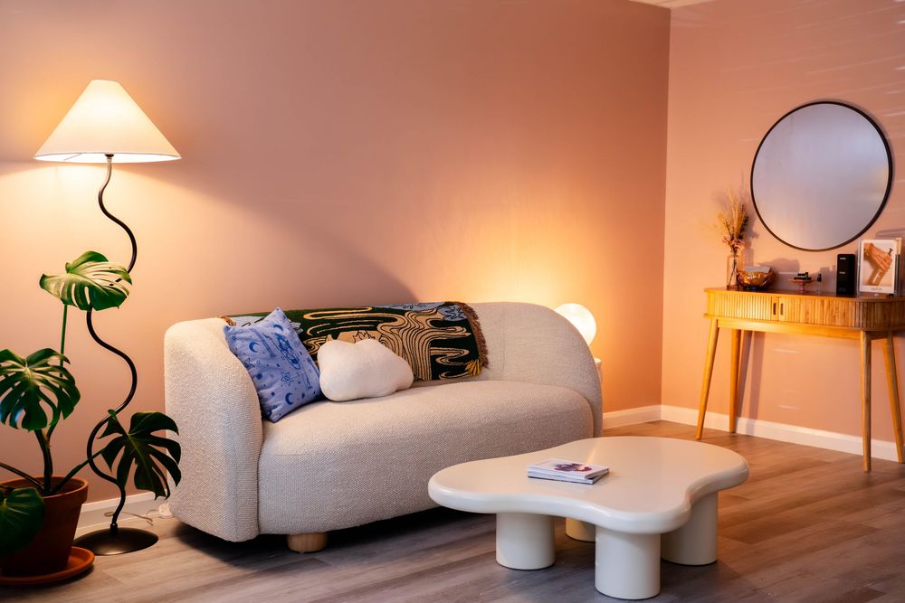 Cozy living room with a beige sofa, round coffee table, floor lamp, and monstera plant against warm pink walls.