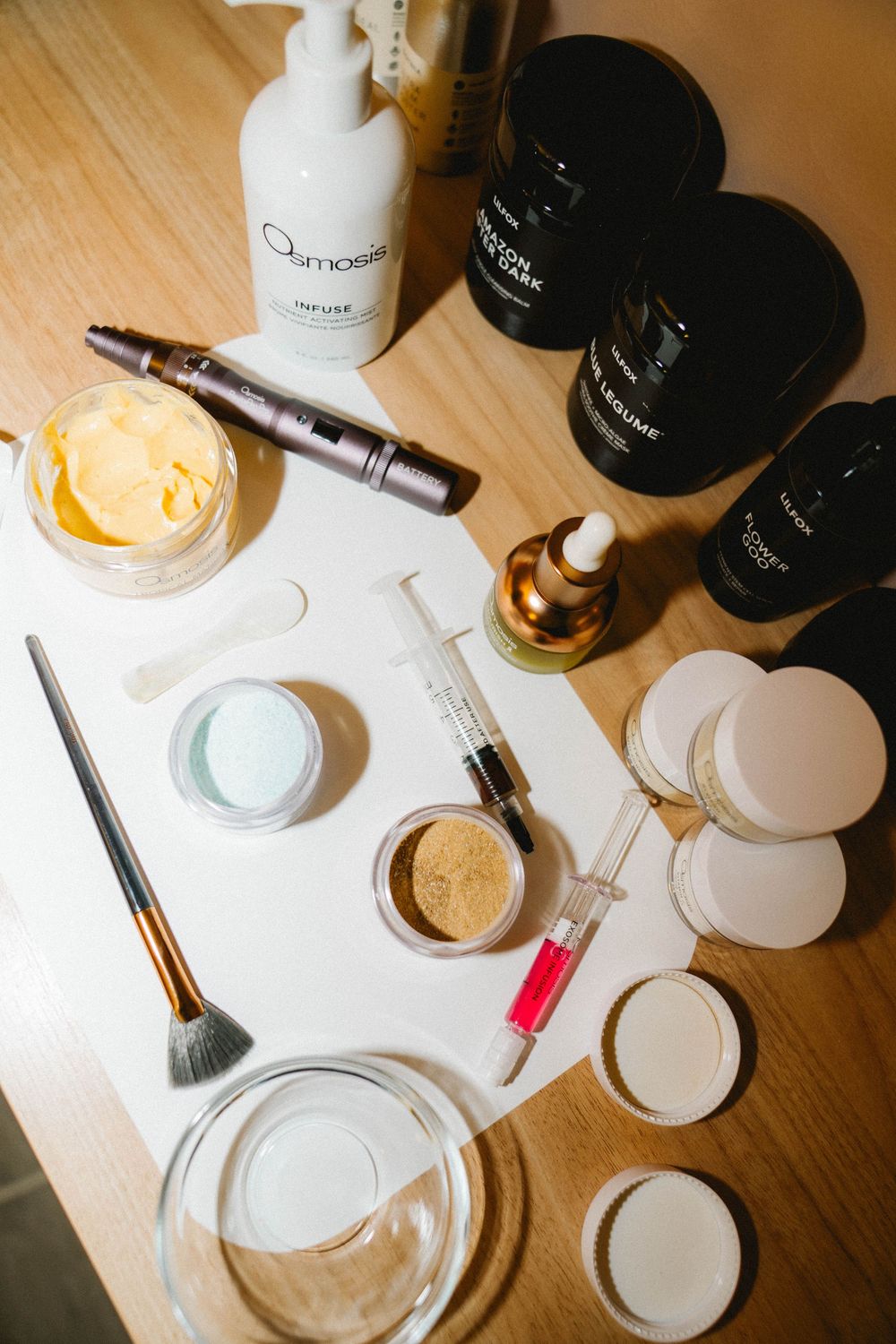 Flatlay of skincare products including Osmosis serum, brushes, syringes, and creams arranged on a white surface.