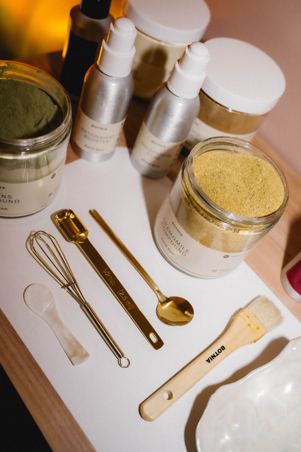 Gold skincare tools and beauty products arranged on a white surface, including spoons, whisk, and jars of powder.