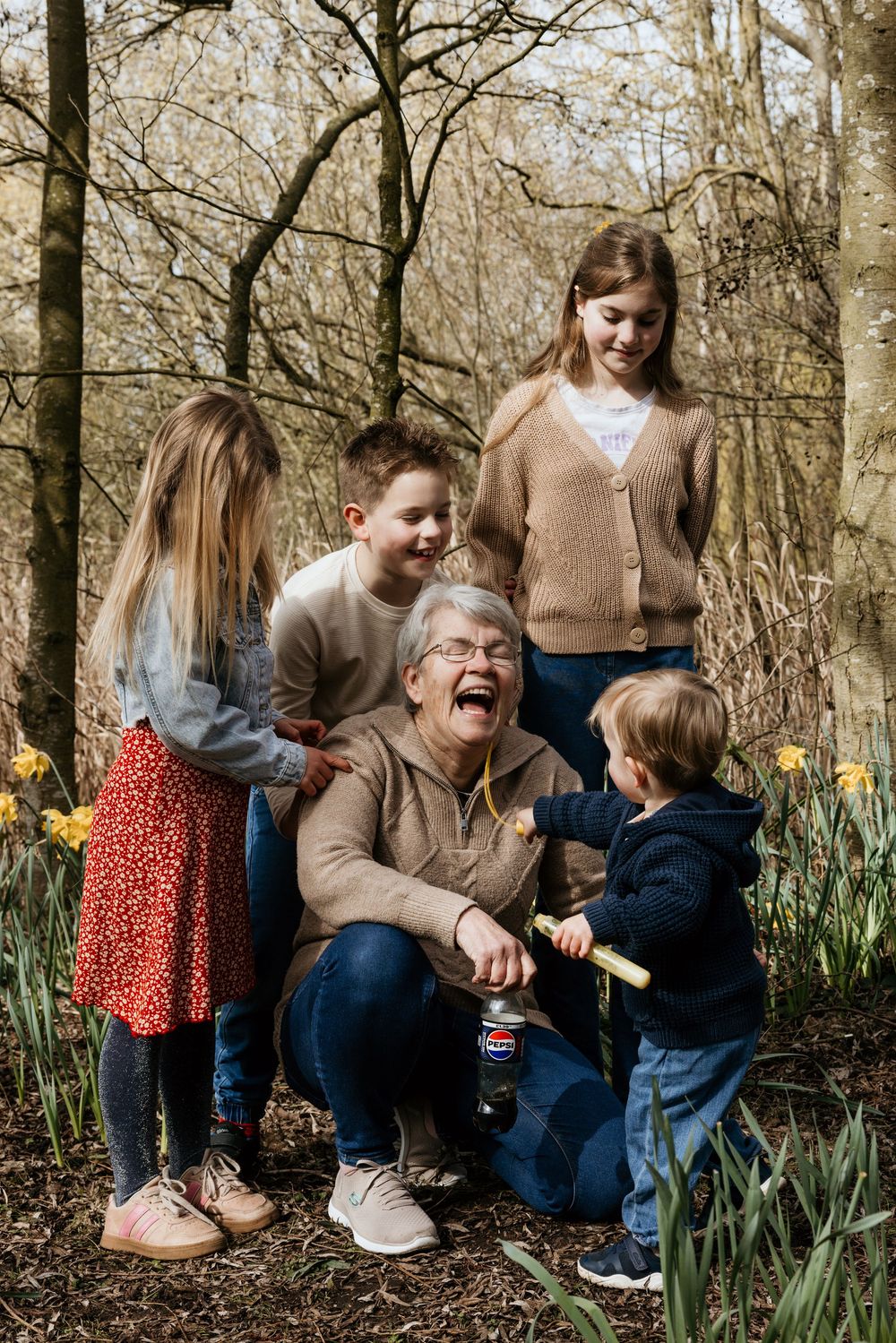 Grandmother laughing joyfully with four grandchildren among daffodils in a woodland setting.