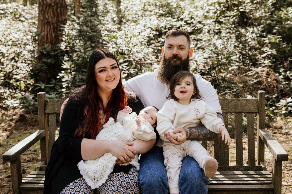 Family of four poses on wooden bench outdoors, parents holding two young children amid lush green foliage.