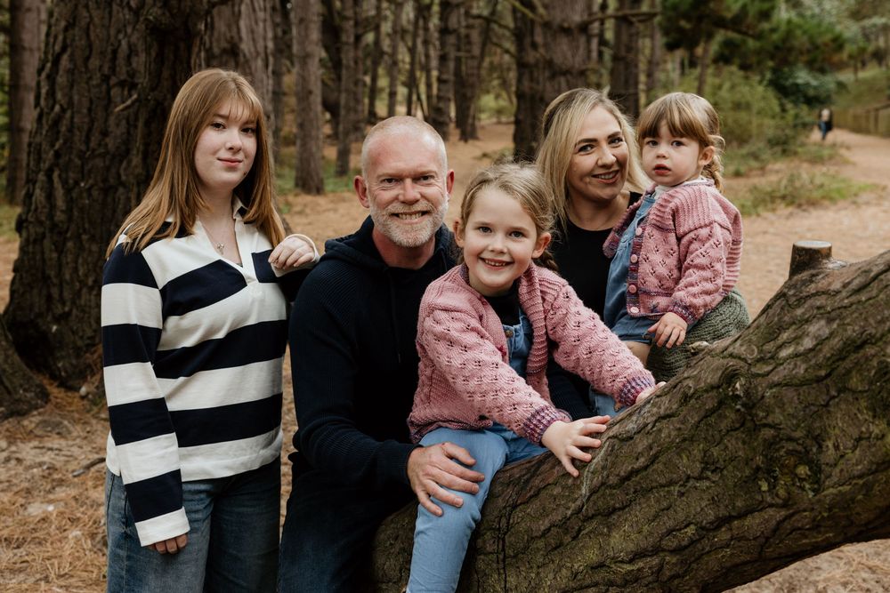 Family of five smiling together outdoors in a pine forest, posing near a fallen log for a portrait.