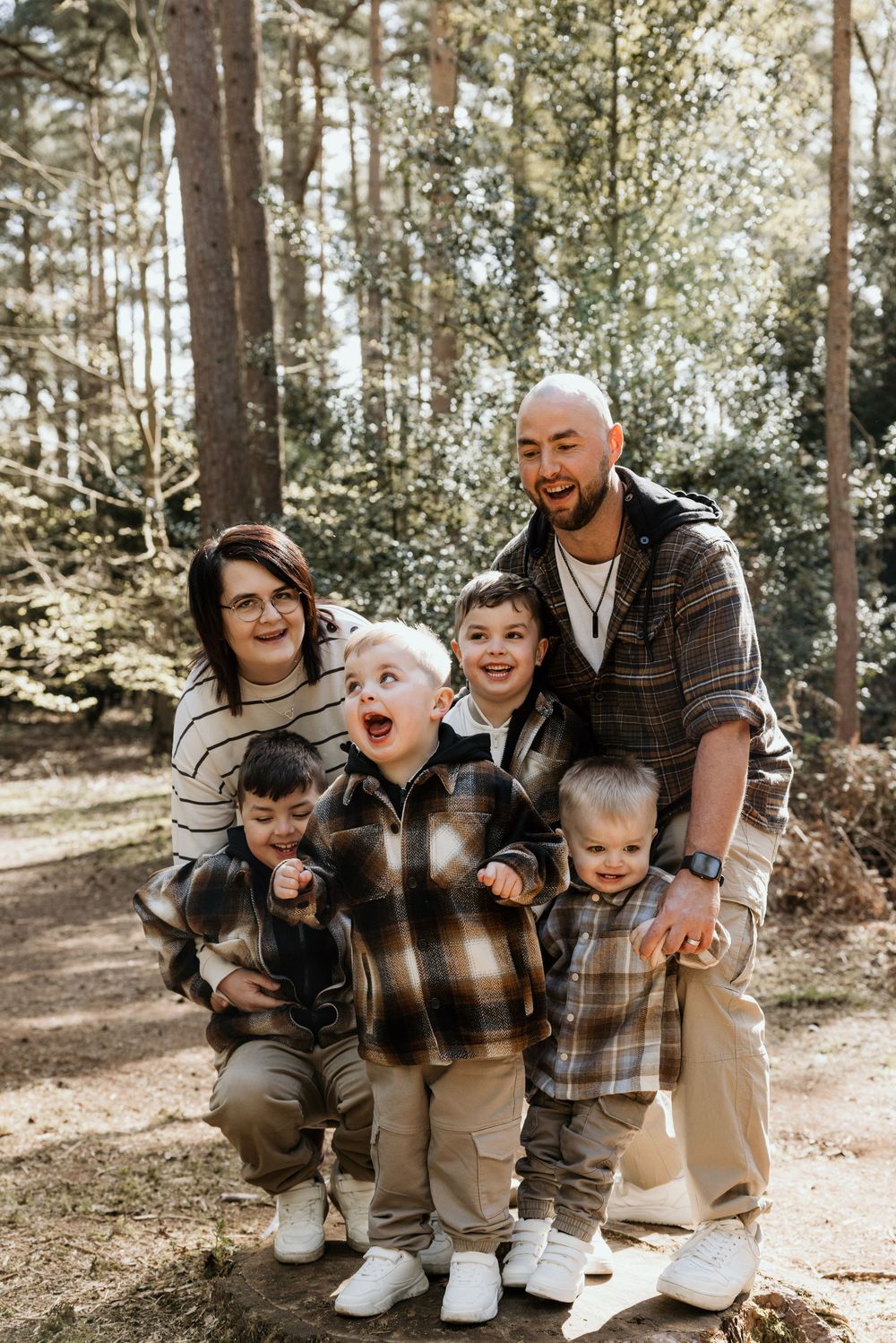 Family of five posing outdoors in a forest, parents with three young boys wearing matching plaid shirts, smiling.