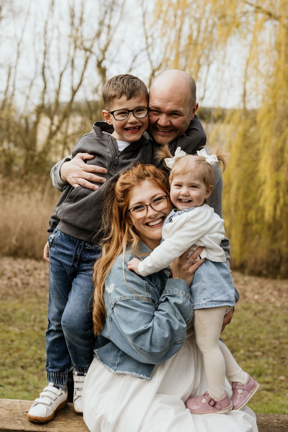 Happy family of four posing outdoors in autumn, parents hugging two young children, smiling warmly in casual attire.