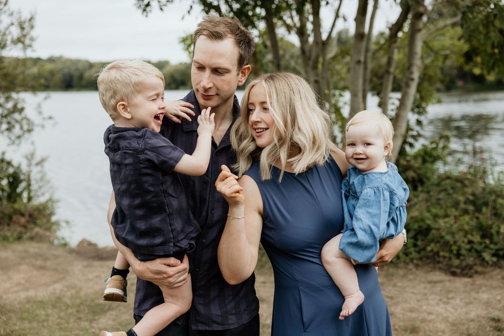 Happy family with two toddlers smiling outdoors near a lake surrounded by green trees.