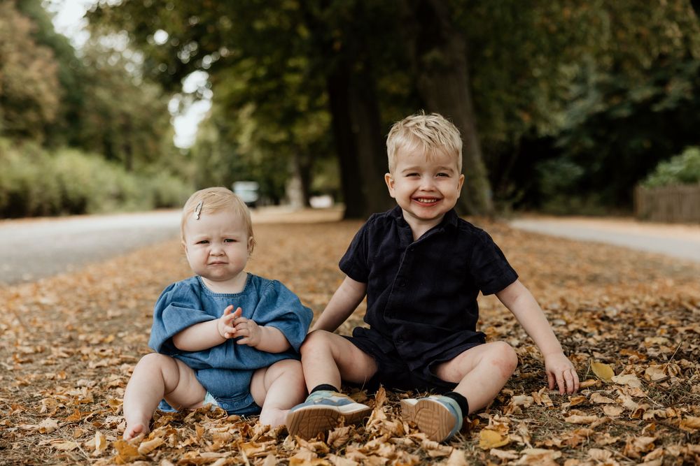 Two young siblings sit smiling among autumn leaves on a tree-lined path.