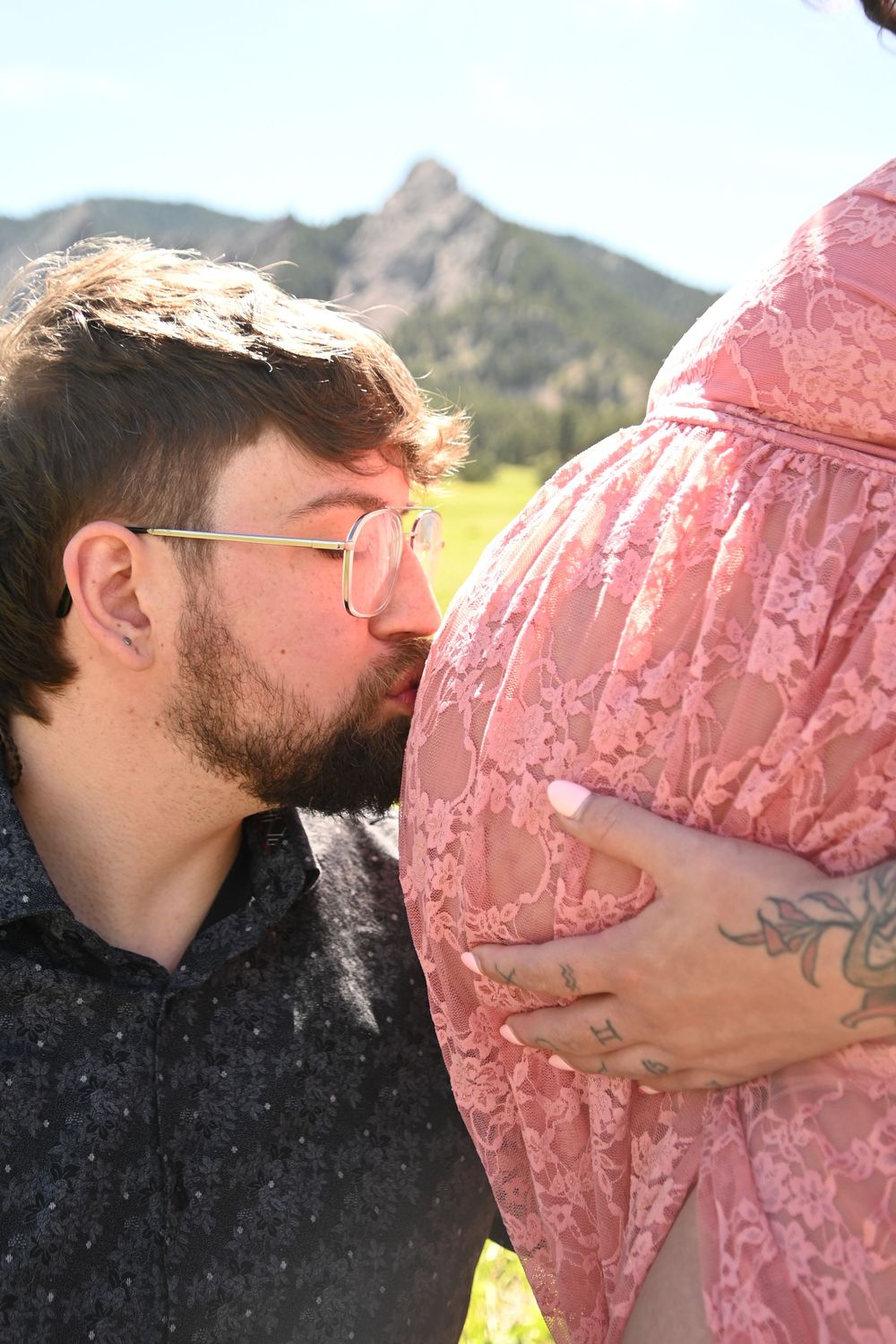 Close-up of an expectant parent kissing pregnant belly in pink lace dress with mountain backdrop.