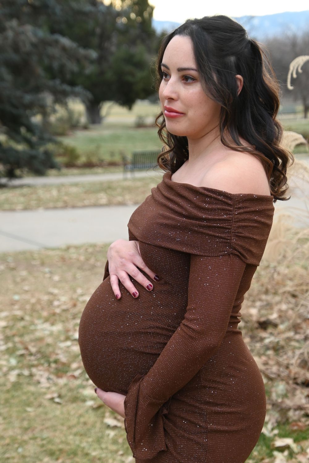 A woman in a brown off-shoulder dress cradles her baby bump while posing outdoors in an autumn setting.