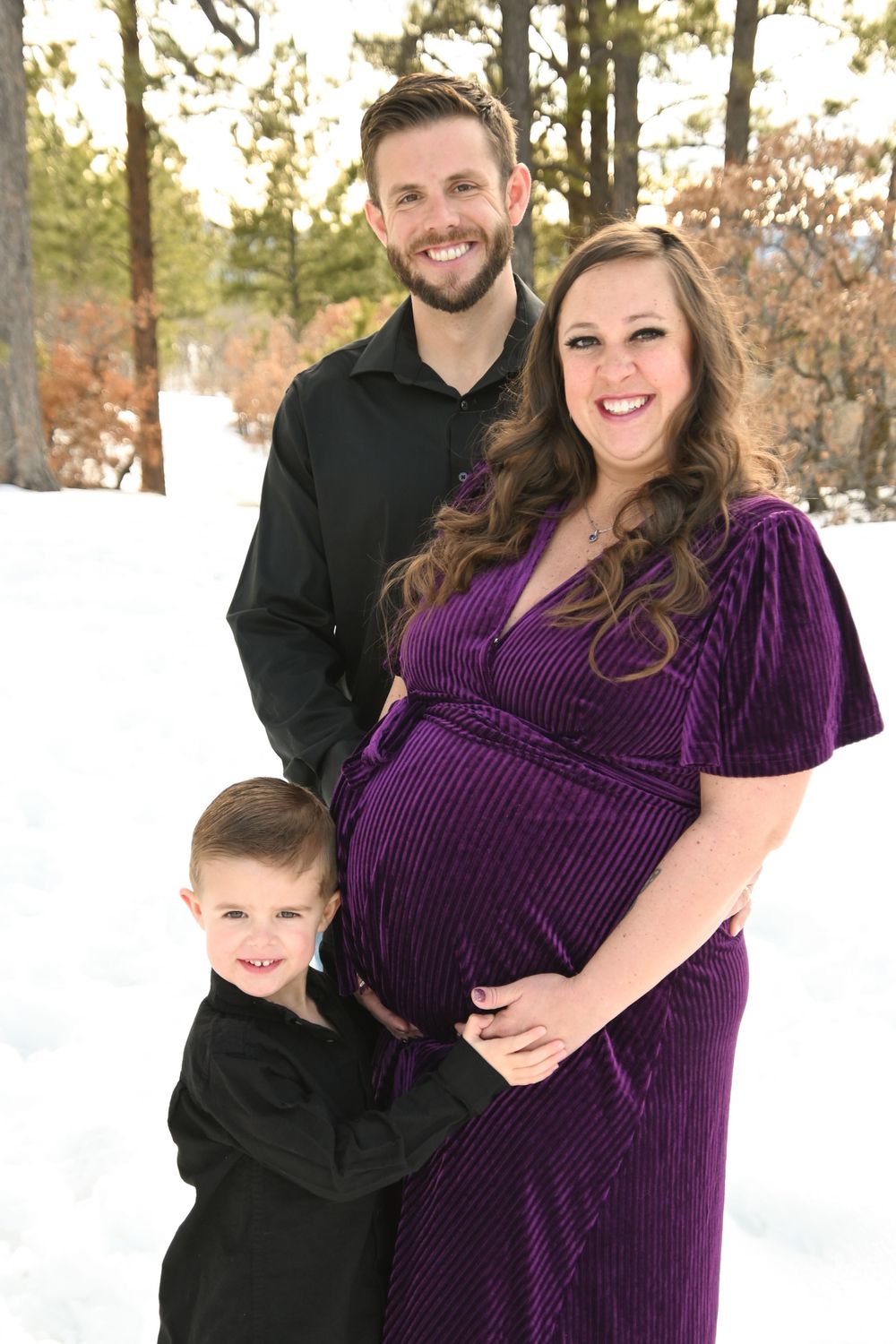 Couple poses for winter engagement photos in snow with woman wearing purple velvet dress and man in dark jacket.