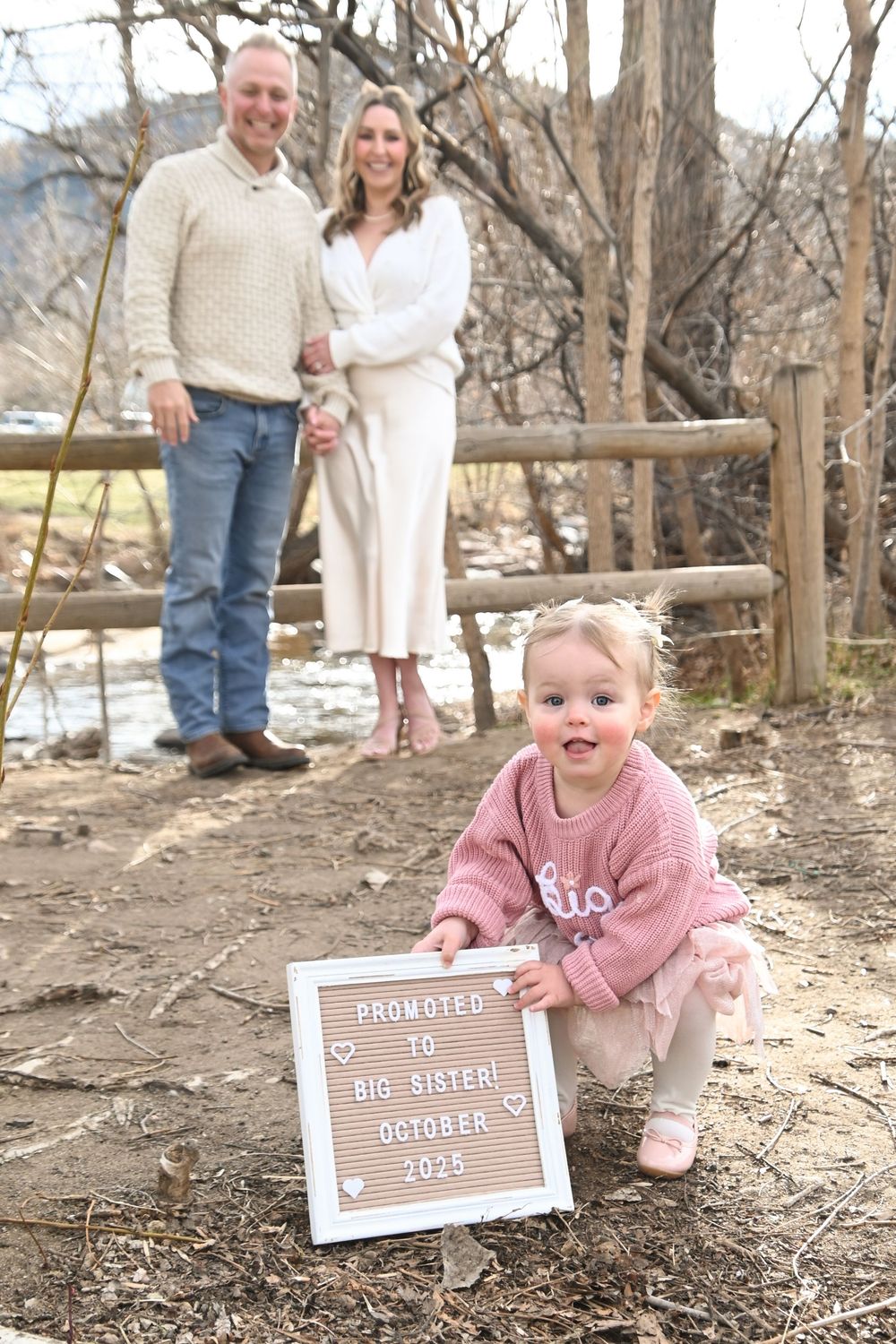 Family poses outdoors near rustic wooden fence for pregnancy announcement with October due date sign.