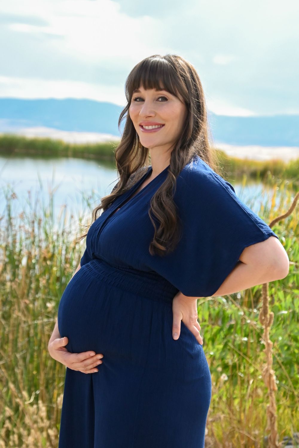 A person in a navy blue dress poses outdoors against a backdrop of tall grass and blue sky during a photo shoot.