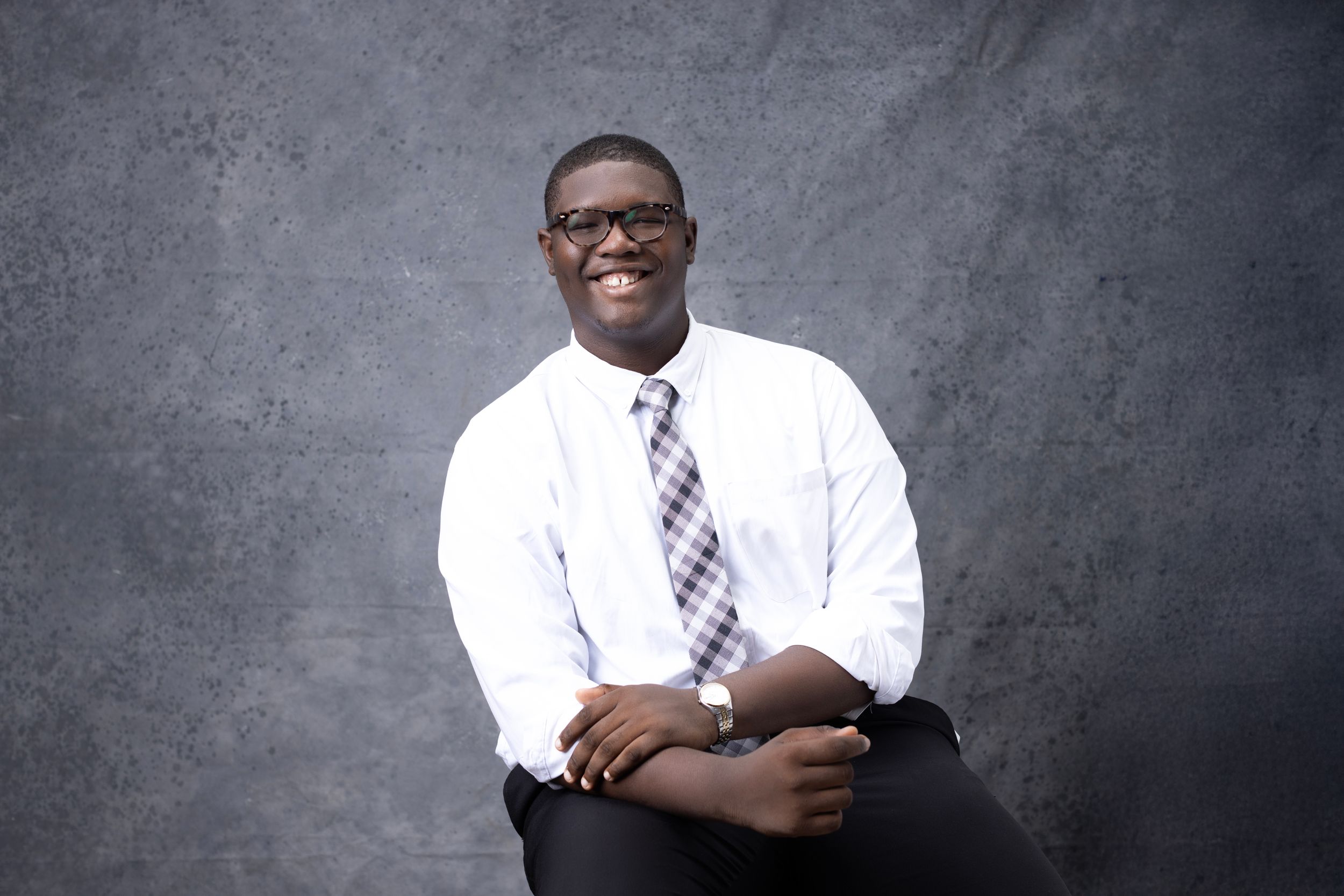 A person wearing glasses, a white dress shirt and purple striped tie smiles while seated against a gray background.