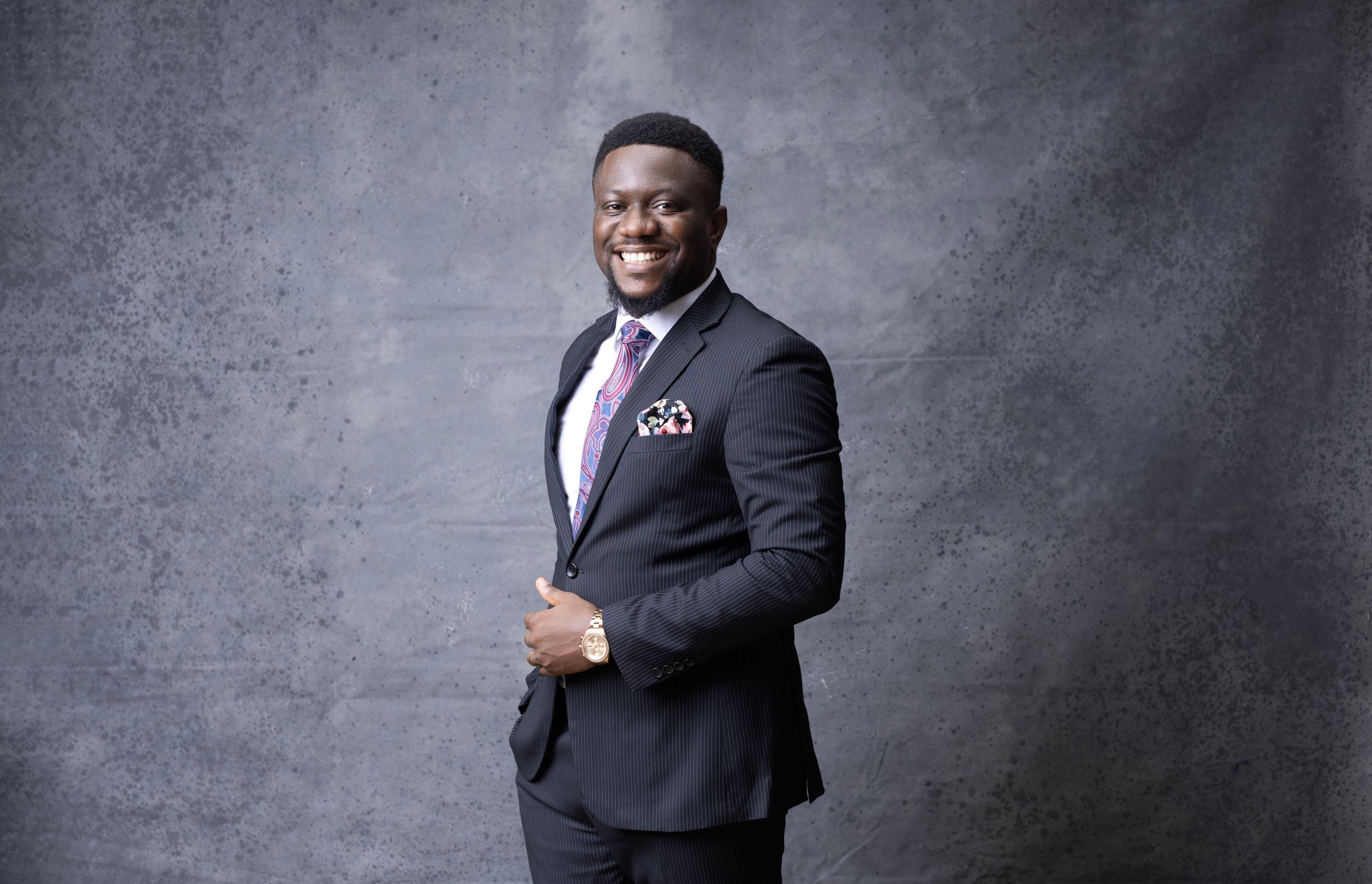 Confident pose in dark suit with pocket square against gray backdrop in studio setting.