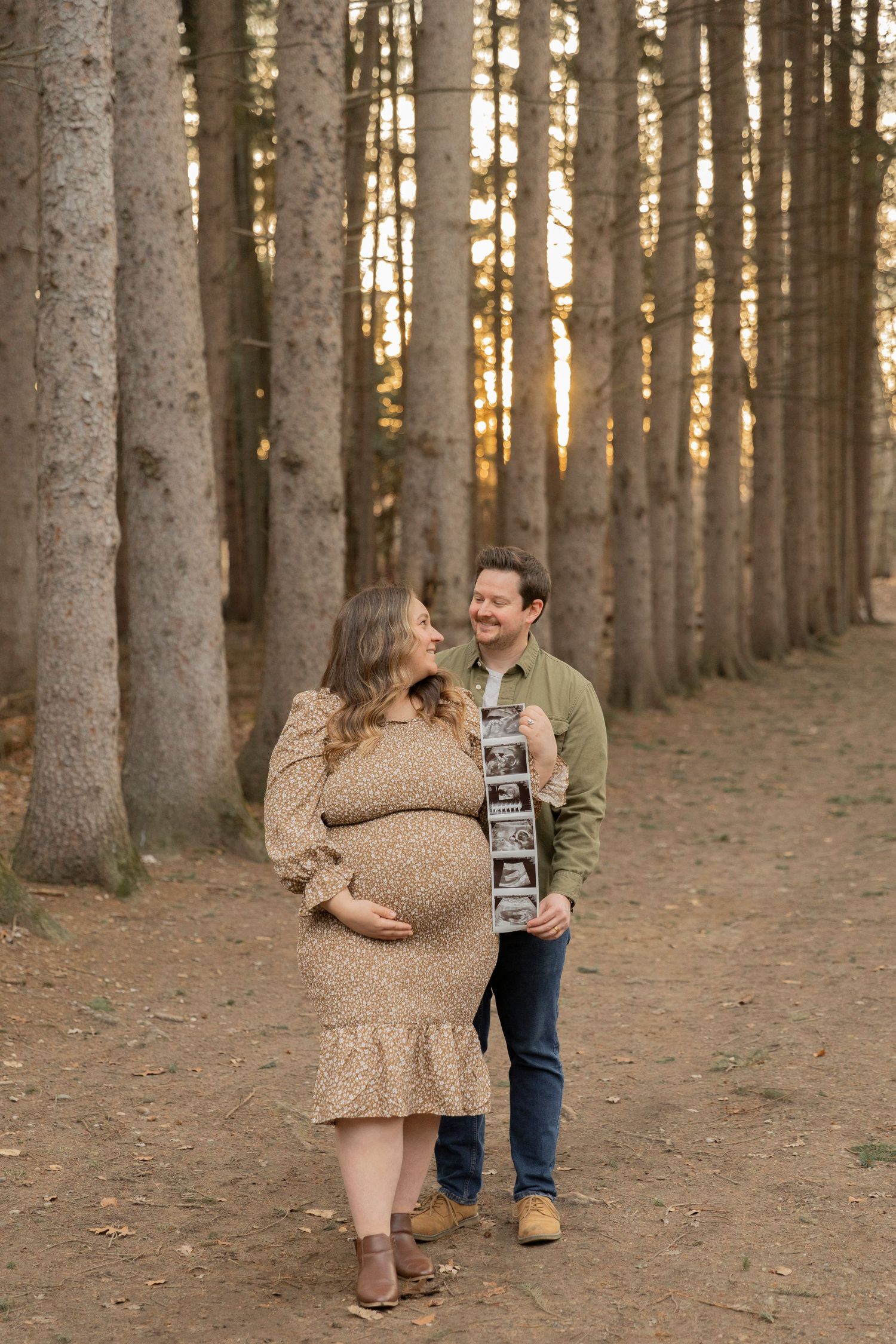 pregnant couple posing at tree line and holding an ultrasound of their baby boy