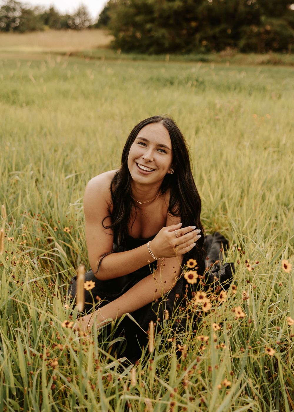 A person in a black dress poses playfully in a golden field of tall grass and wildflowers during sunset.