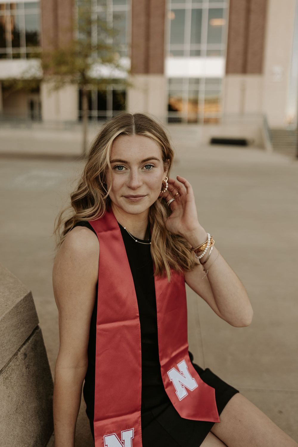 Graduate wearing a red graduation stola over black dress poses outside university building in warm lighting.
