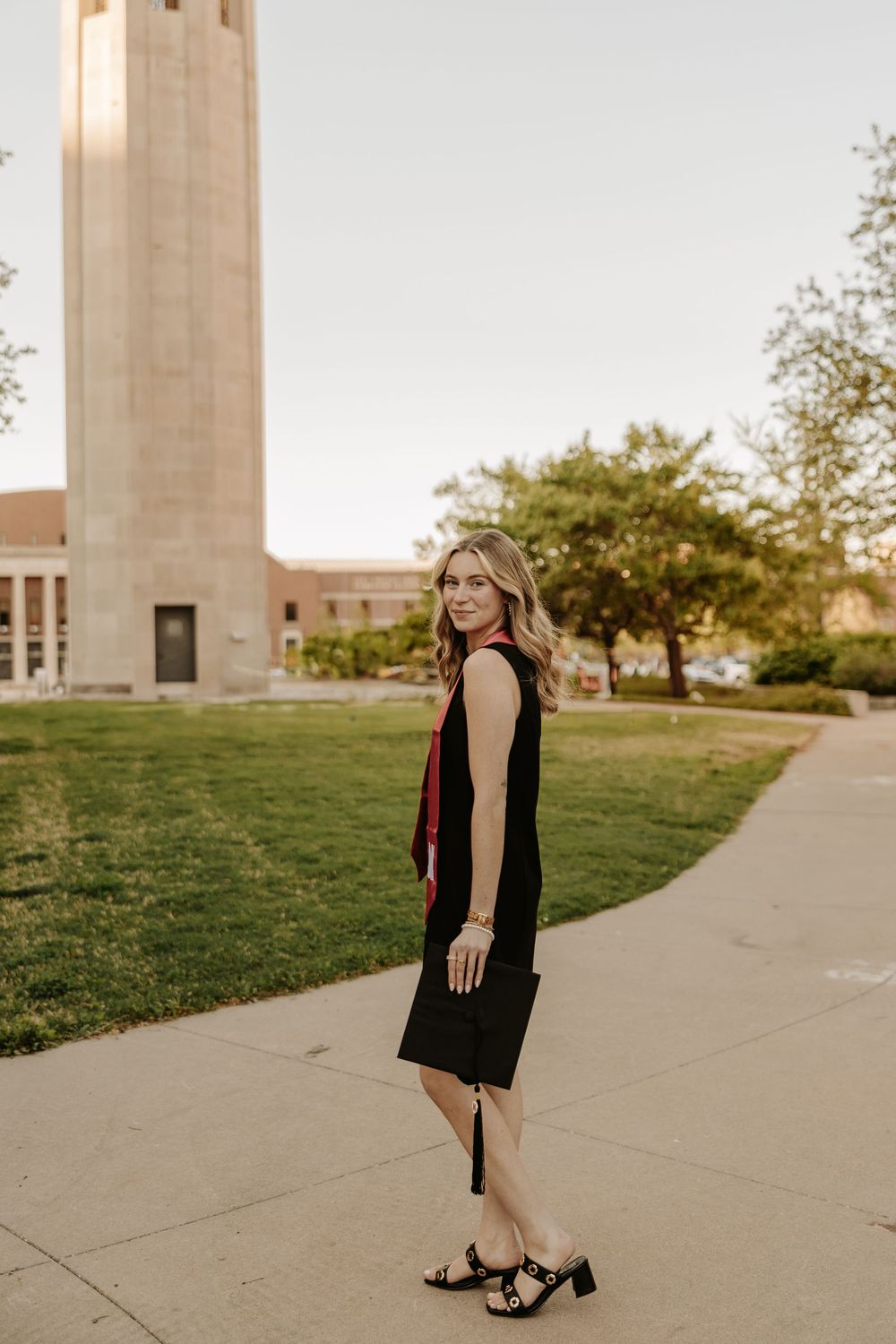 Graduate in black dress stands on pathway with tall university bell tower visible in warm evening light.