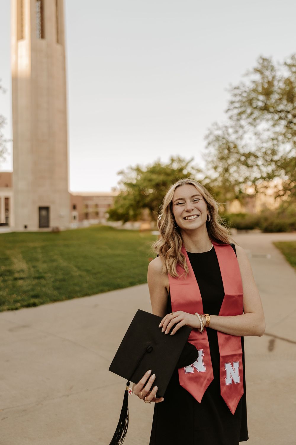 Graduate in a black dress and coral stole celebrates with diploma near university bell tower at sunset.