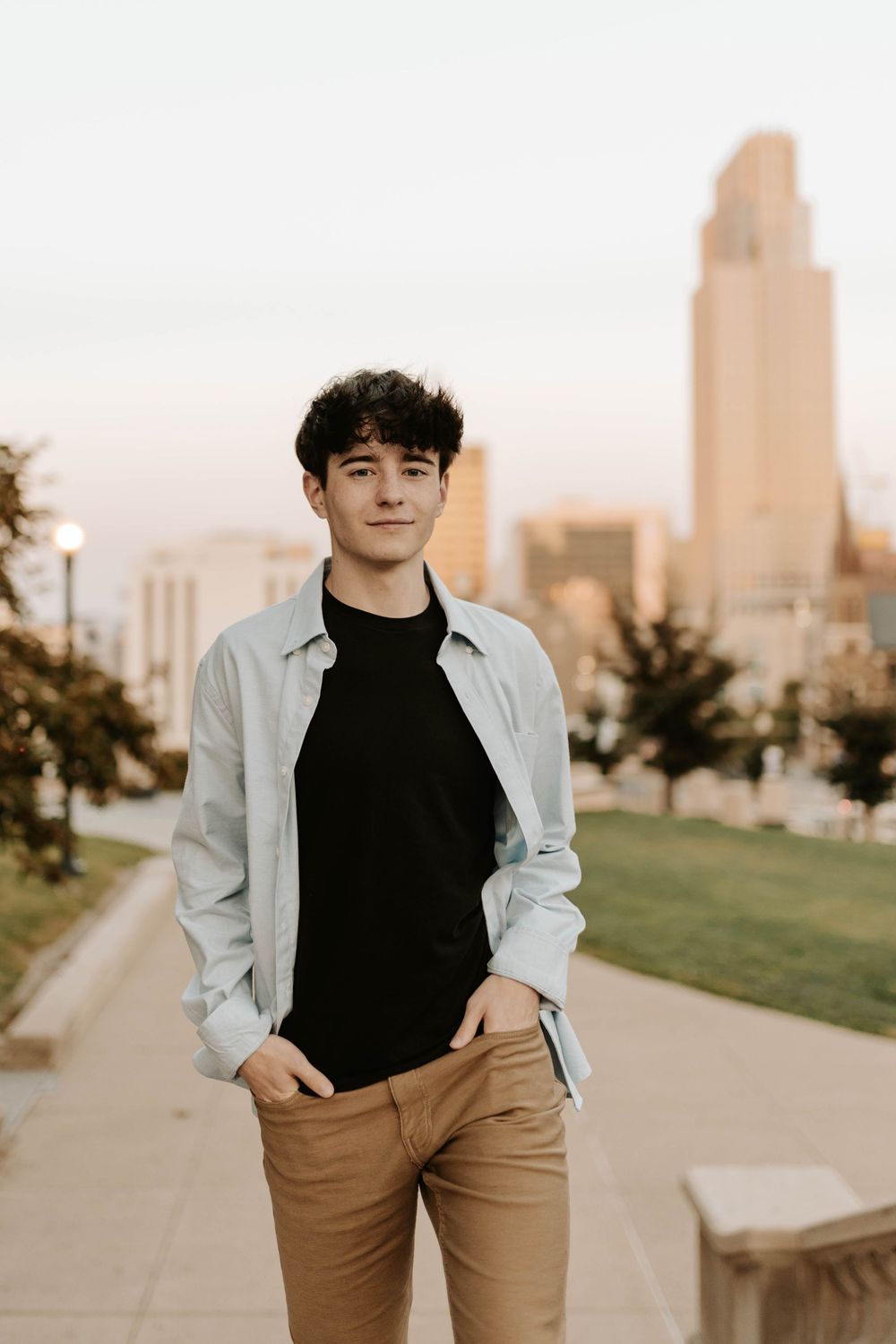 A person in a black turtleneck and light jacket poses outdoors with a city skyline visible in the background.