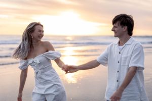 Engaged couple wearing all white shirt and dress on the beach during golden sunset behind holding hands and laughing