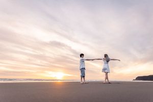Engaged couple wearing all white shirt and dress on the beach, golden sunset behind holding arms spread out against sky