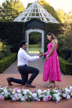 Man one one knee proposes Engagement Ring in Hamilton rose garden pergola at sunset