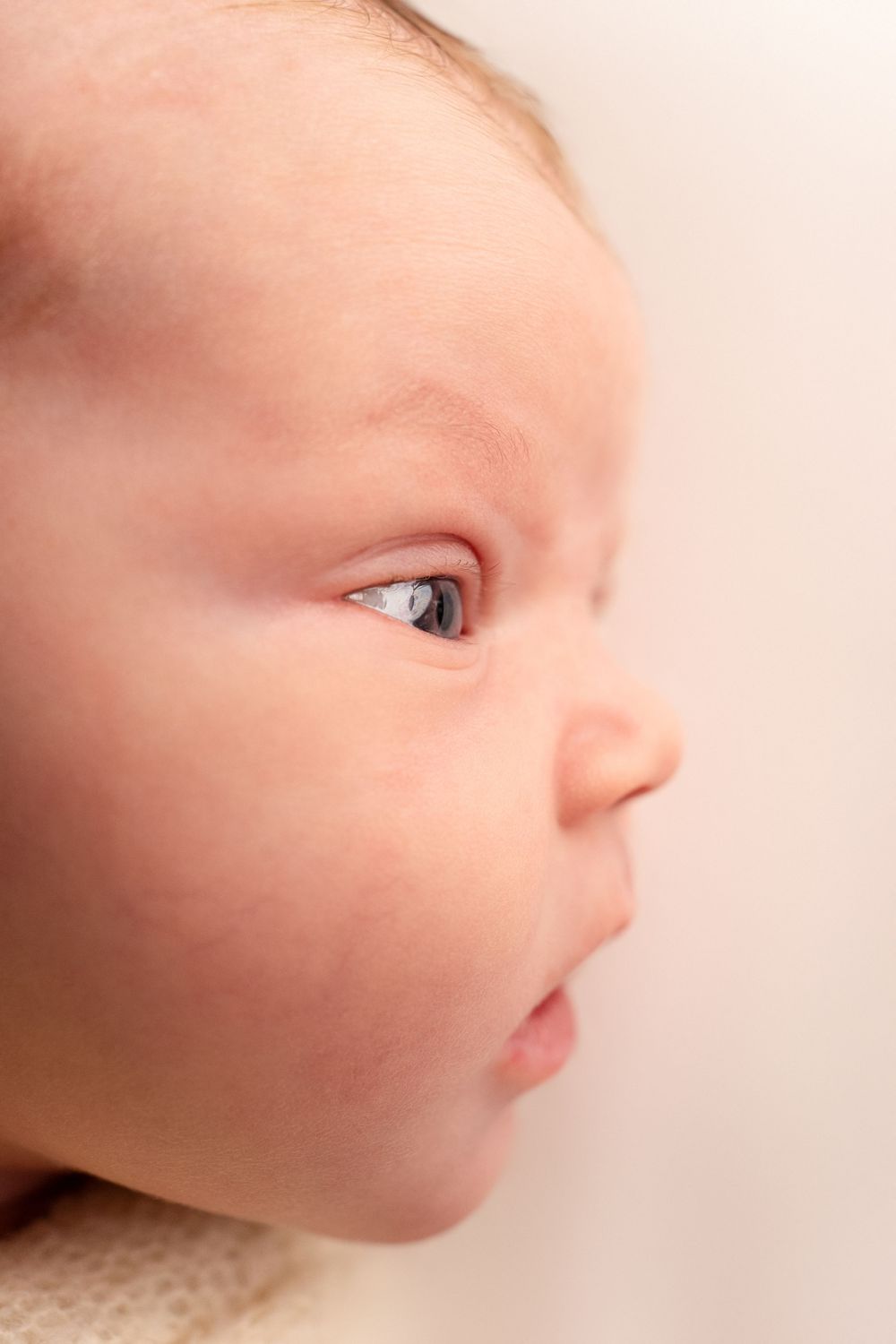 Close up side profile view of a baby with soft skin and bright eyes looking to the side against light background.