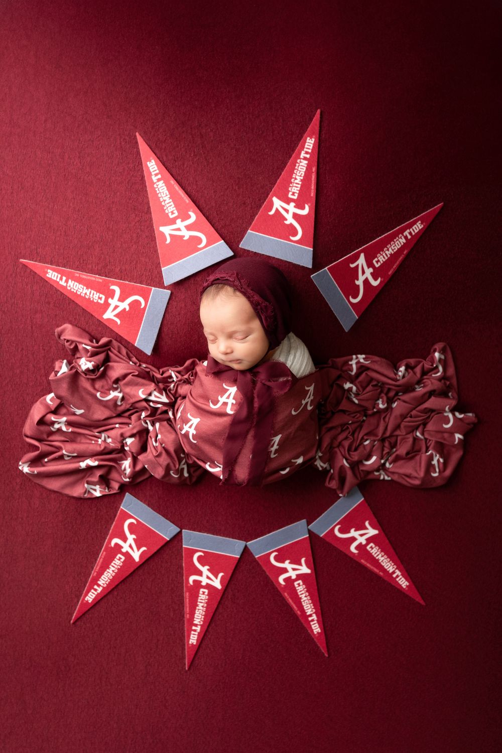 Newborn photography with Alabama Crimson Tide themed setup featuring red pennants arranged in star pattern on crimson background.