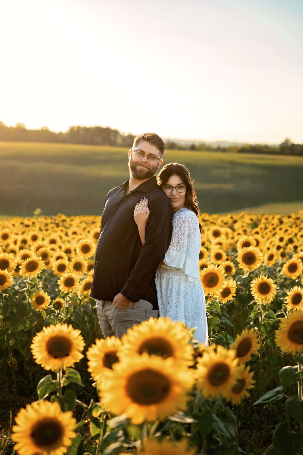A couple embraces in a golden sunflower field at sunset with warm sunlight illuminating the vibrant yellow blooms.