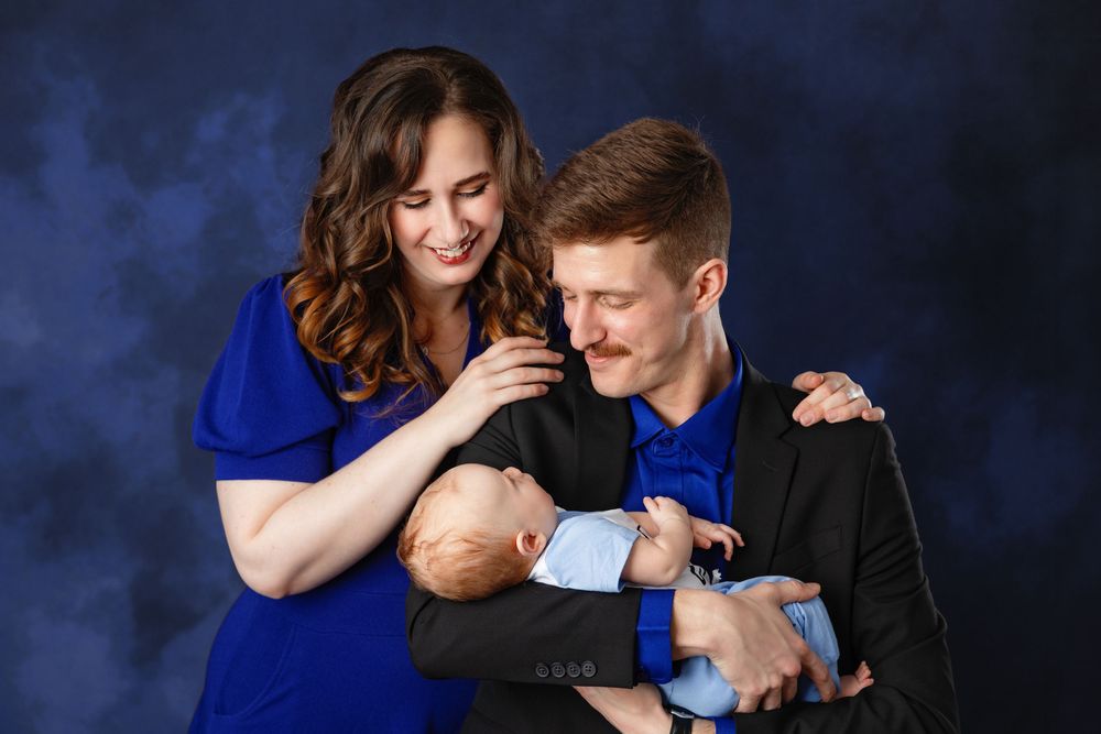 A couple in blue and black outfits share a tender moment with their newborn baby against a dark smoky background.