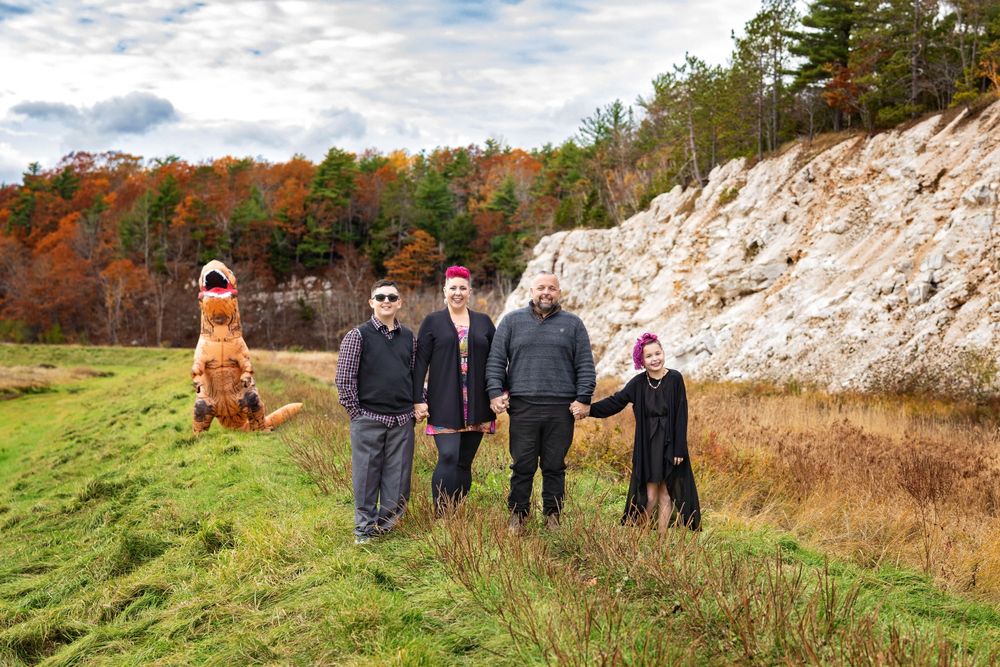 A group outdoors stands near a rocky cliff face with autumn foliage in the background on a sunny day.