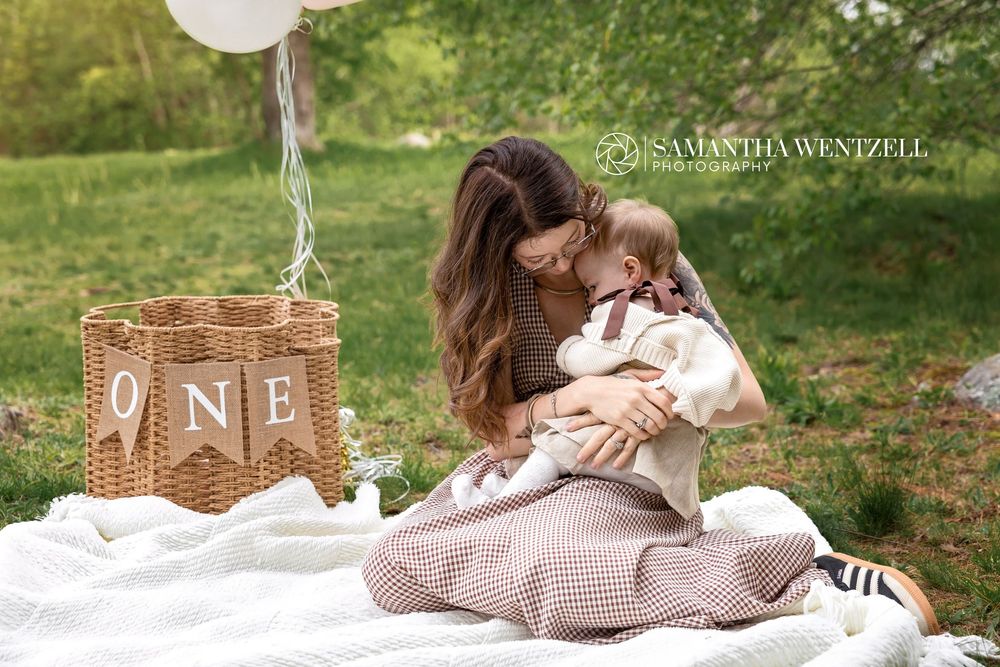 Mother cuddling baby at first birthday picnic with ONE banner and balloon by Samantha Wentzell Photography.