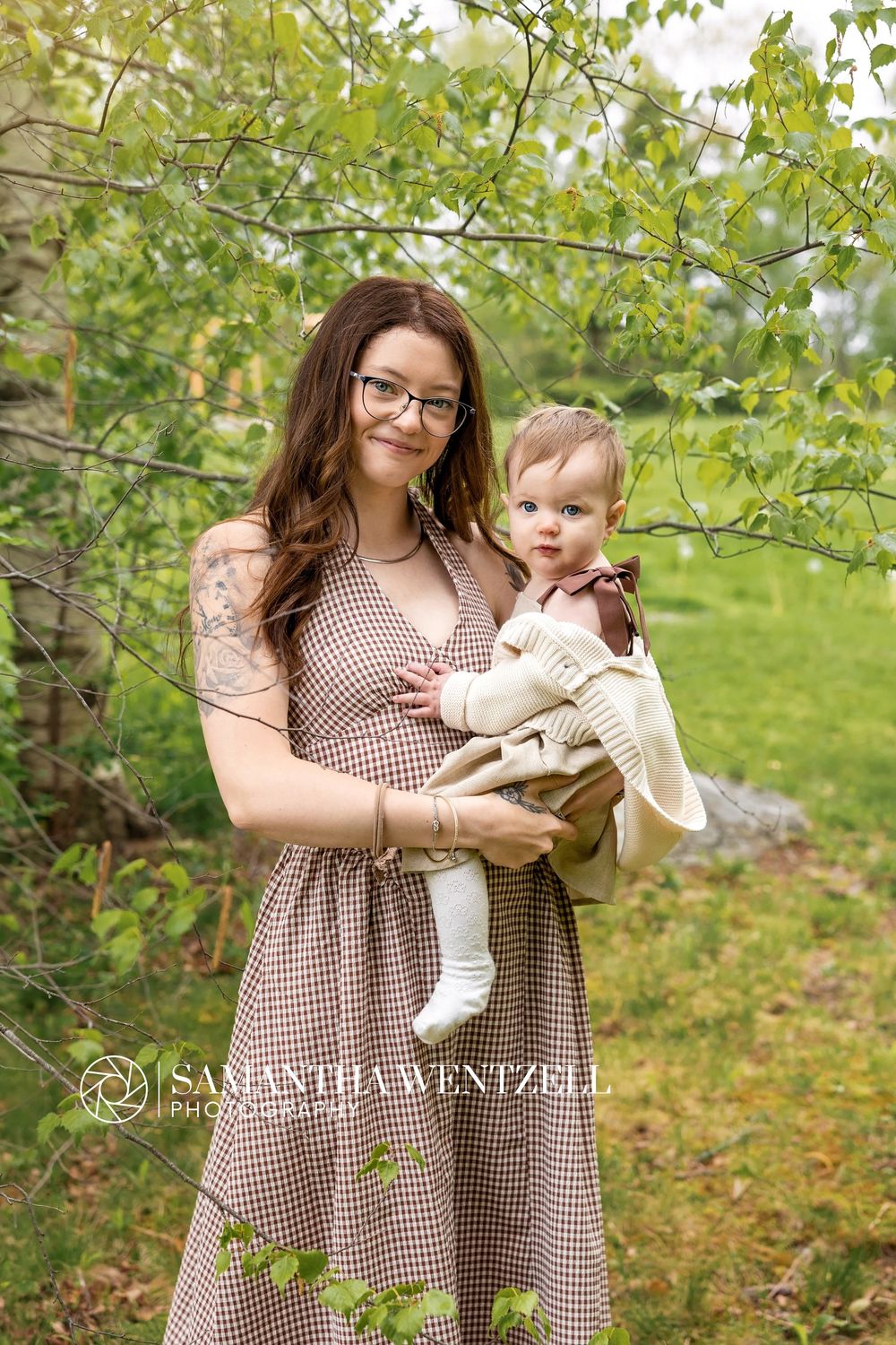 A mother in a brown gingham dress holds her baby outdoors among green trees, photographed by Samantha Wentzell.