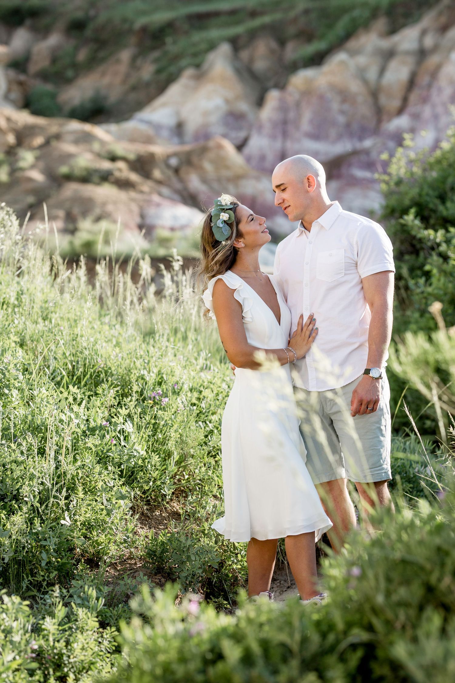 A romantic couple in white attire share an intimate moment among wildflowers with rocky cliffs in the background.