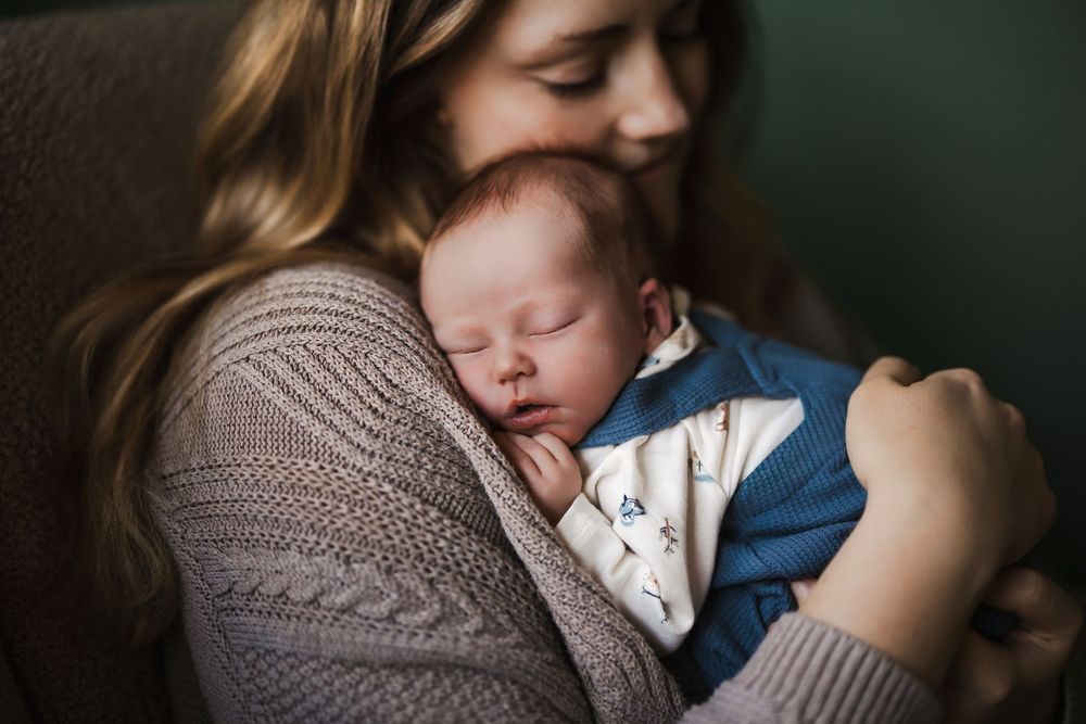 A mother tenderly holds her sleeping newborn baby dressed in blue against her chest.