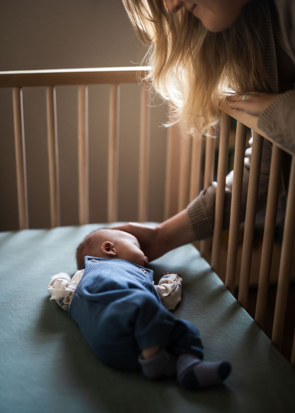 Mother gently placing newborn baby in wooden crib, soft warm light, tender parenting moment.