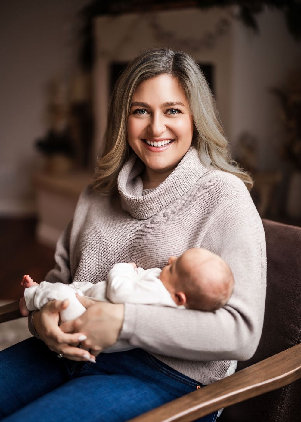 Smiling woman in beige turtleneck sweater holding a newborn baby while sitting in a wooden chair.