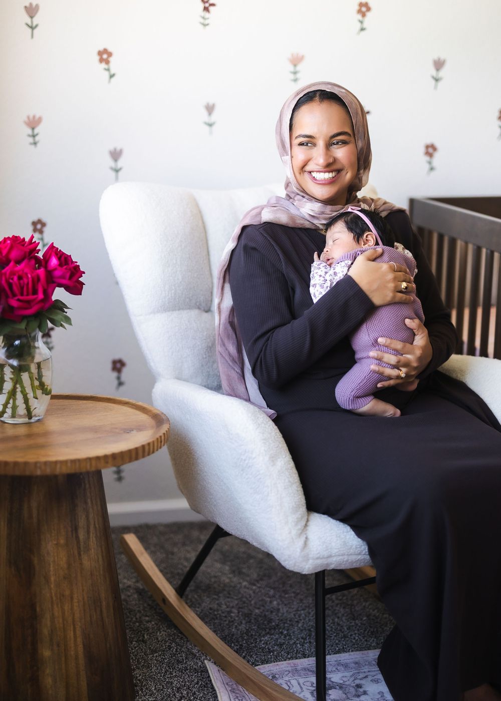 A person in a black dress and hijab sits comfortably in a white armchair next to a wooden side table with flowers.