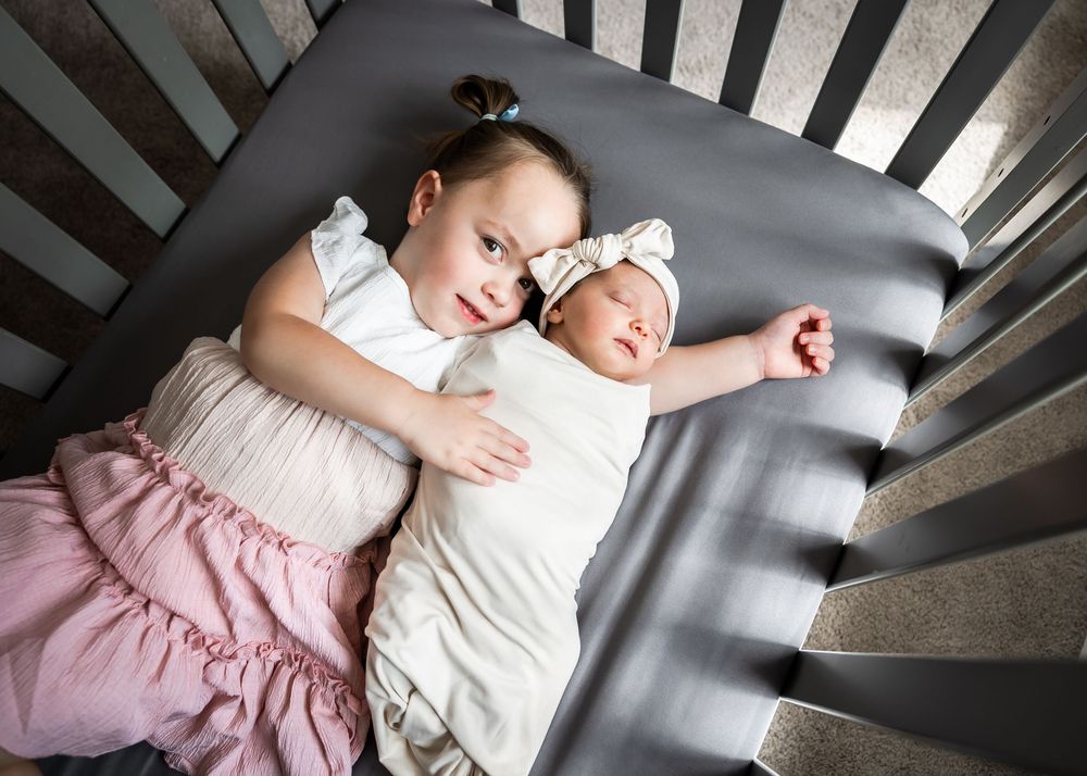 Two infants sleeping peacefully side by side in a wooden crib with grey bedding.