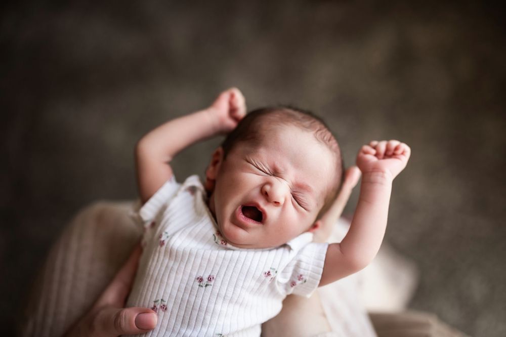 Newborn baby yawning and stretching arms while being held, wearing a white floral onesie against a gray background.