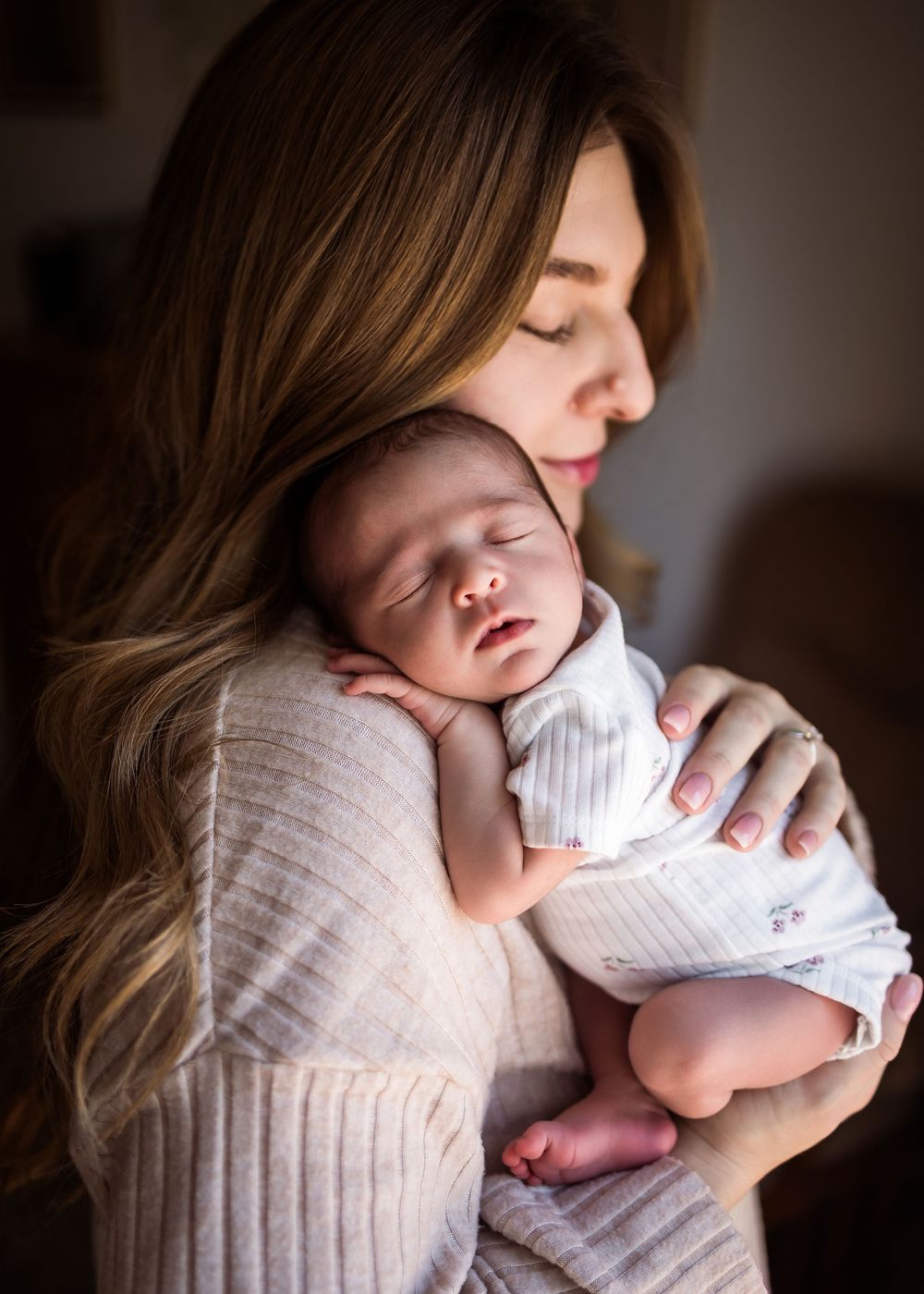 A mother tenderly holds her sleeping newborn baby against her chest, both with eyes closed in a warm, soft-lit room.