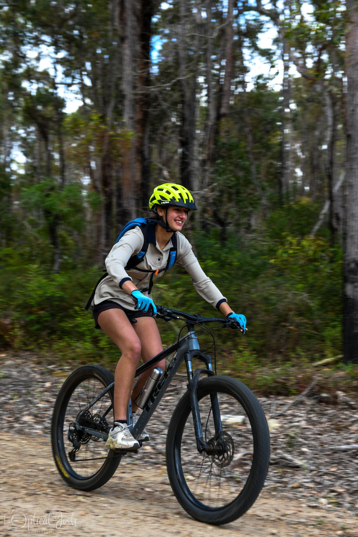 Mountain biker rides a trail through a forest wearing protective gear and neon yellow helmet. (Optical Josh)