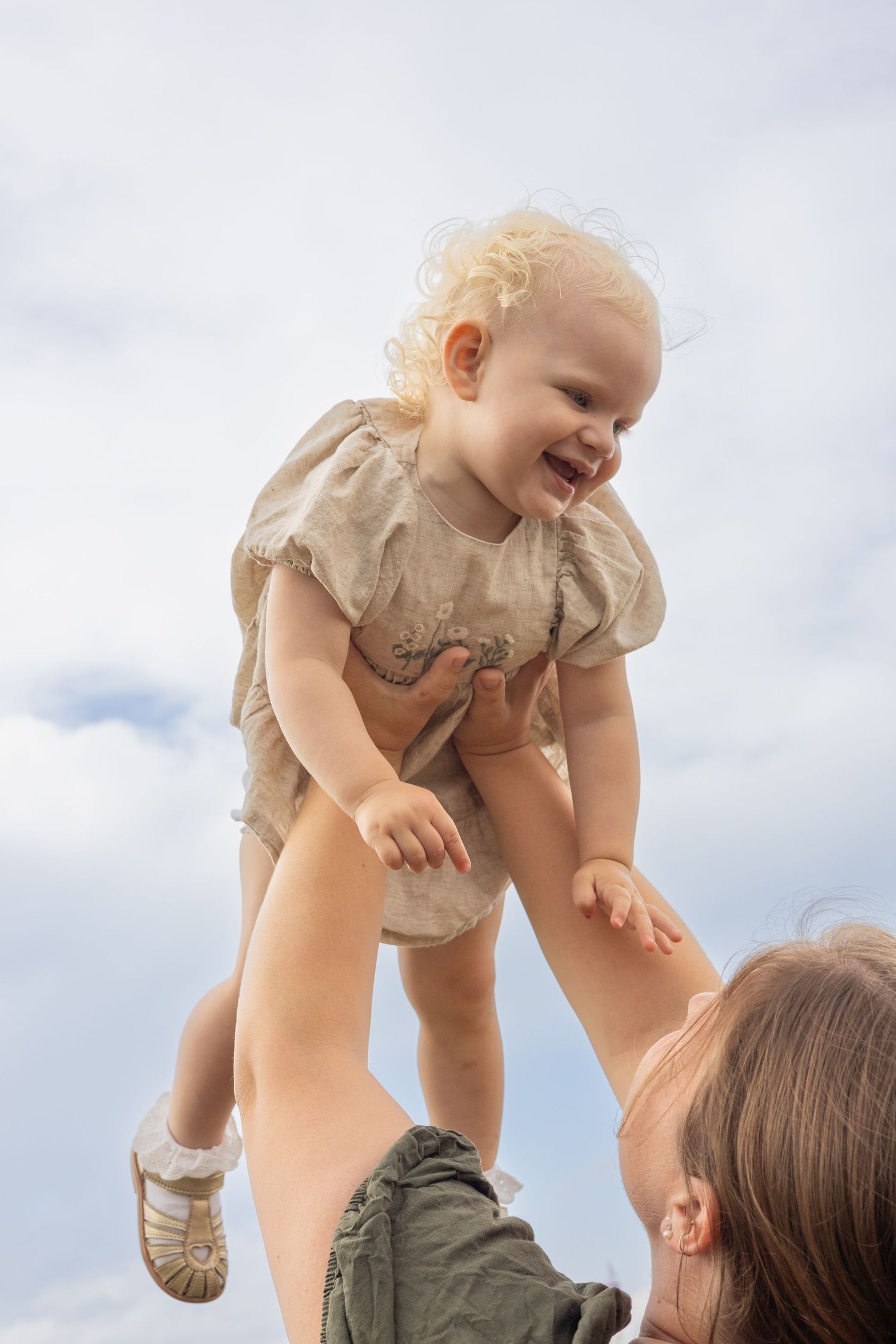 Parent lifts laughing toddler in beige dress against cloudy sky.