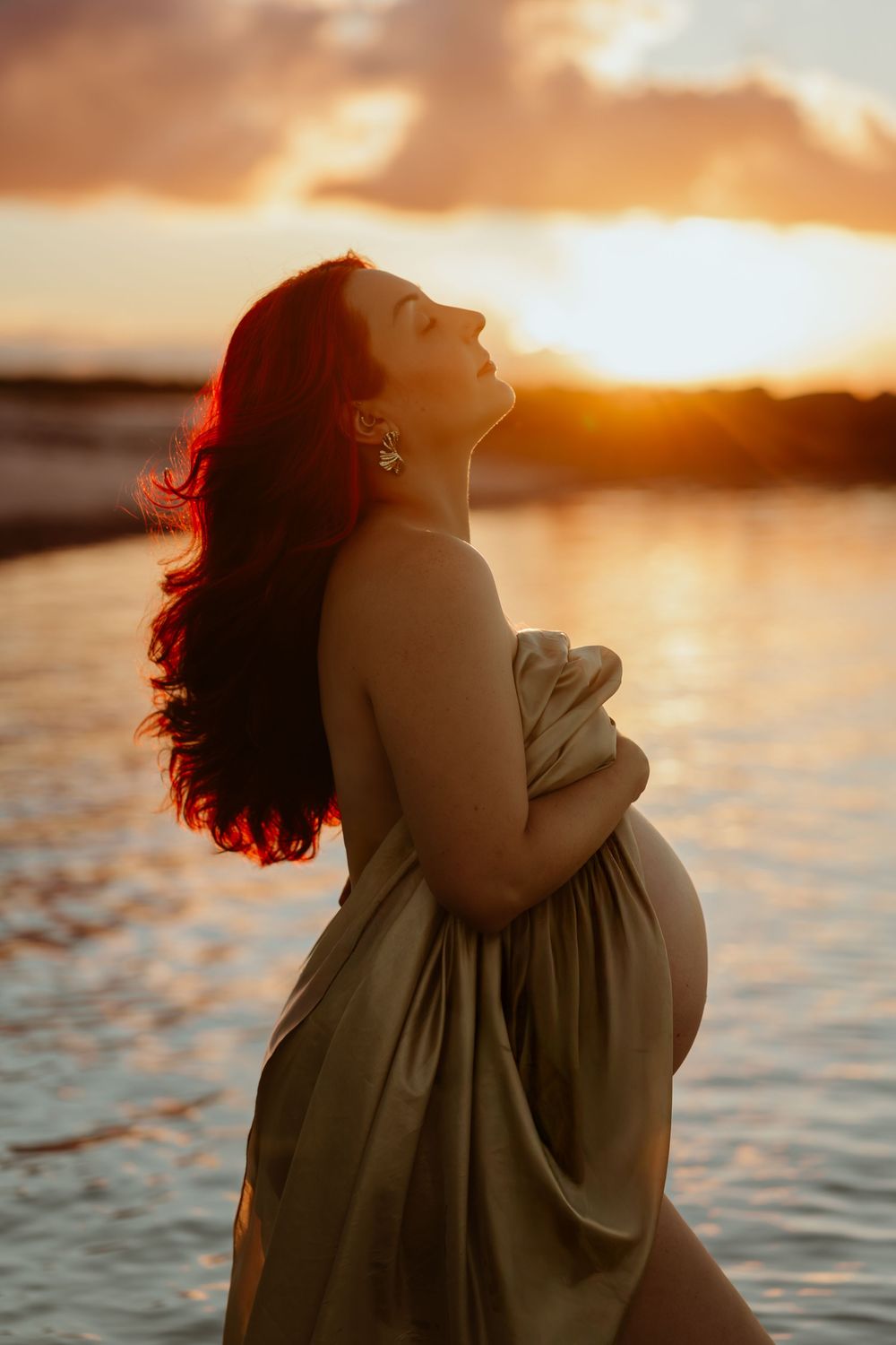 Pregnant woman wrapped in golden fabric poses gracefully at sunset beach, glowing light illuminating her silhouette.