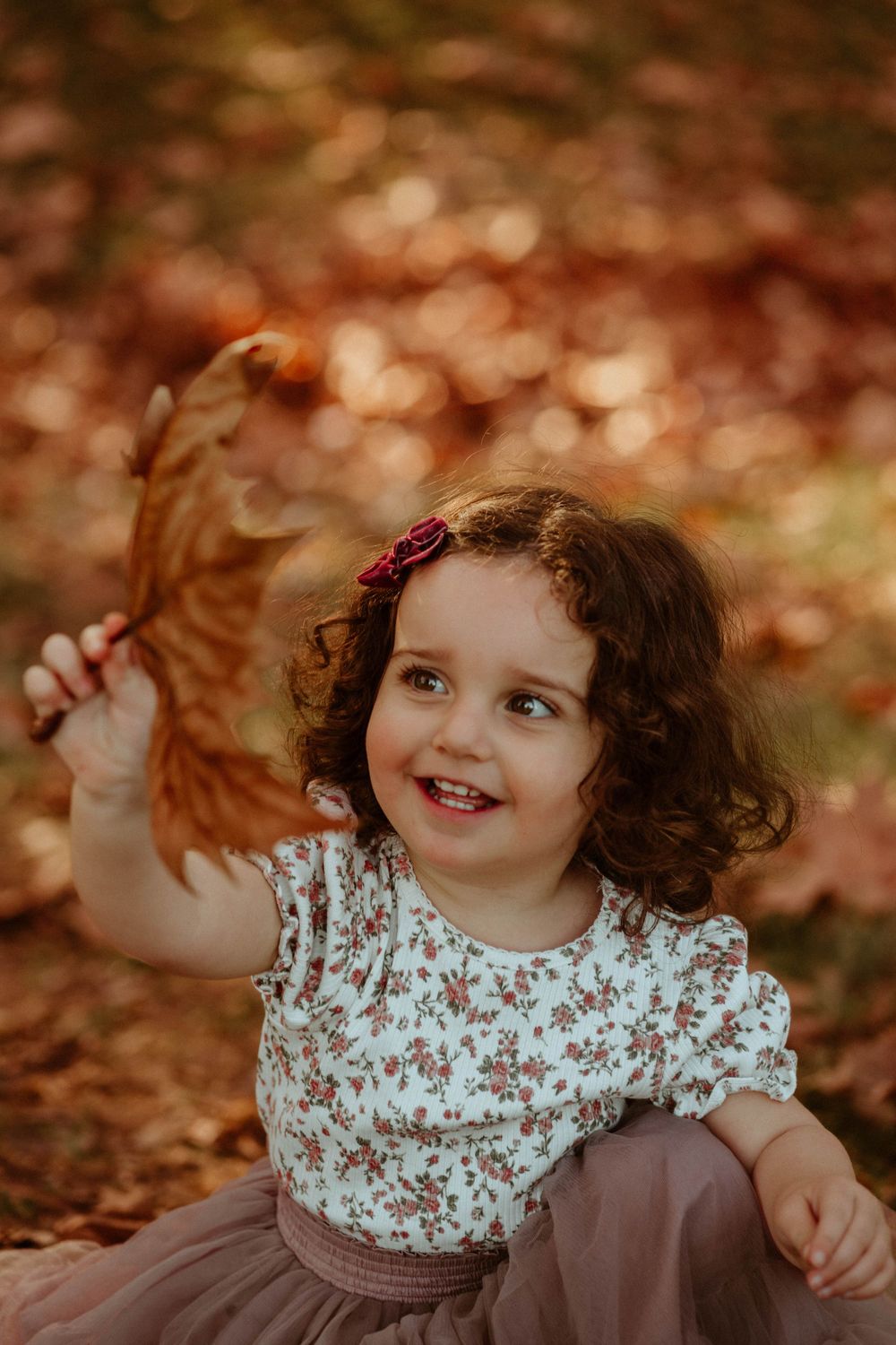 Curly-haired child smiling while playing with autumn leaves.