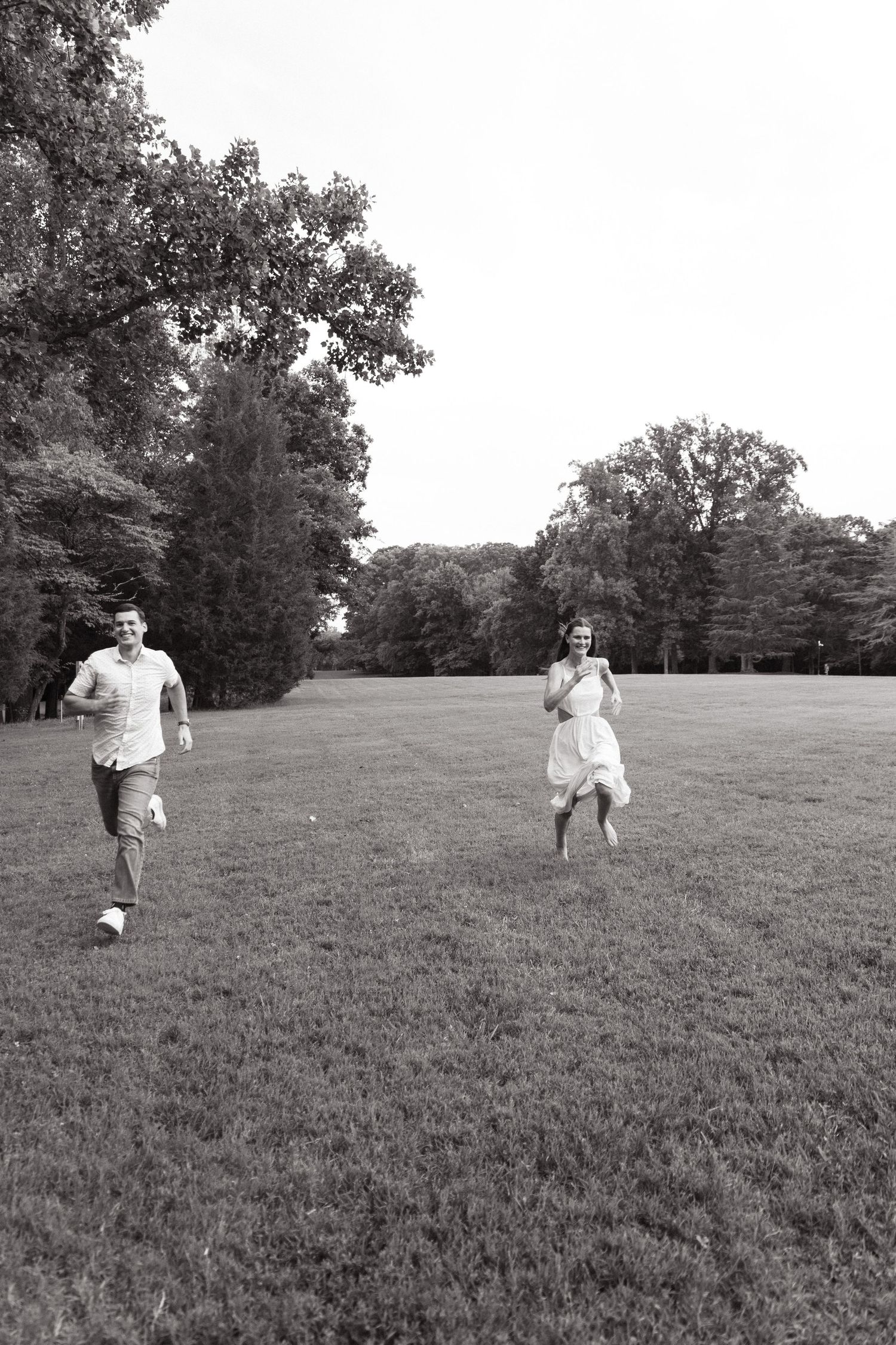 Couple running playfully across an open grassy field in black and white.