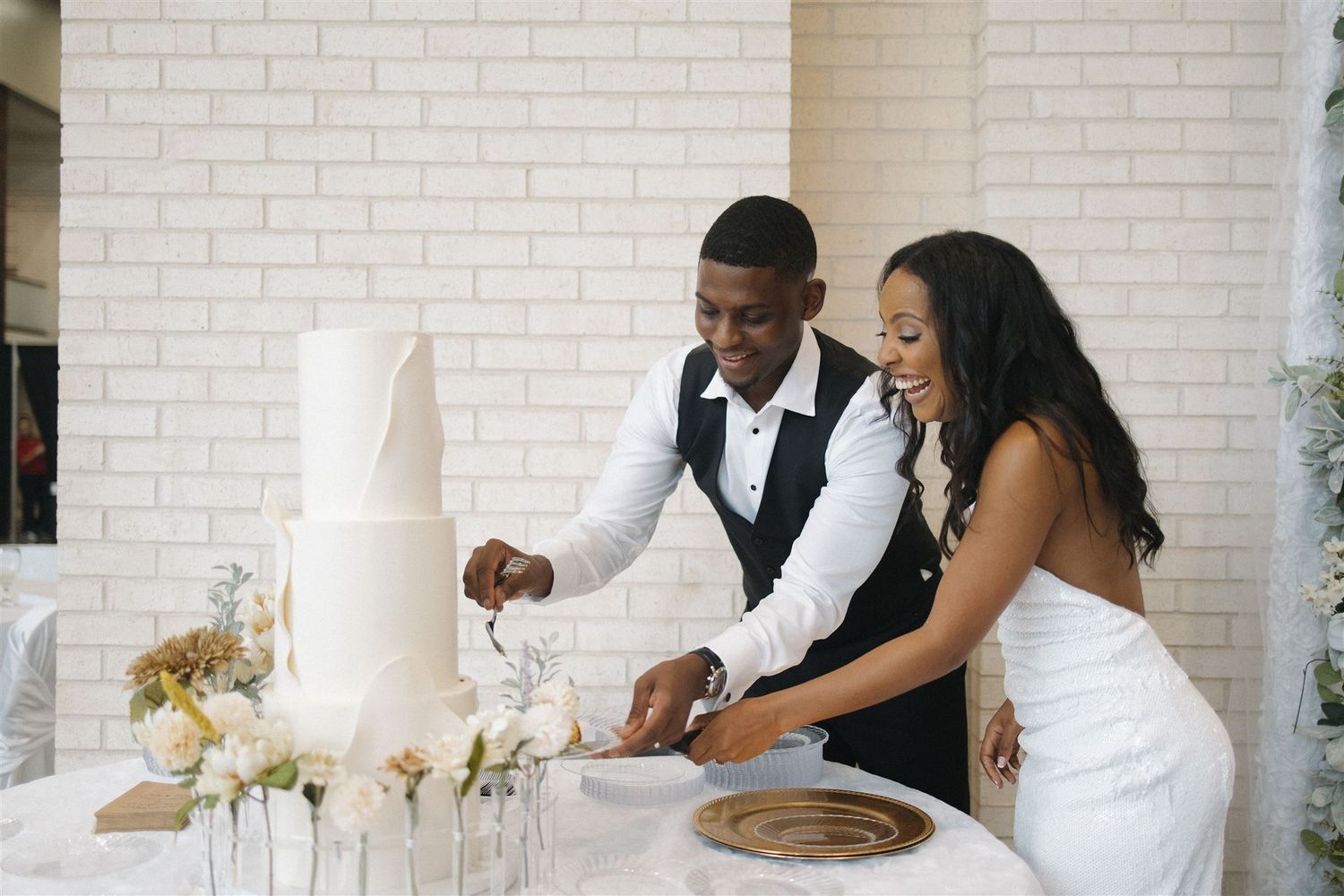 A couple in formal wedding attire joyfully cut into their elegant white wedding cake together.