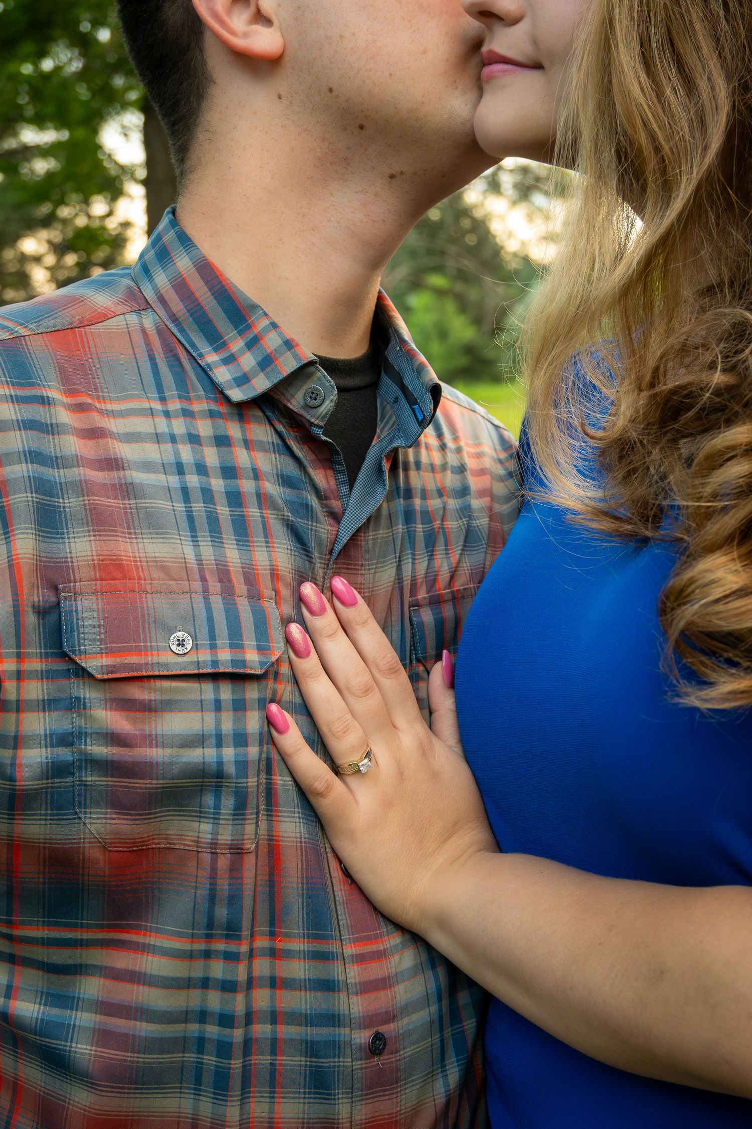 A newly engaged couple smiling for the camera. She has her hand on his chest showing her ring.