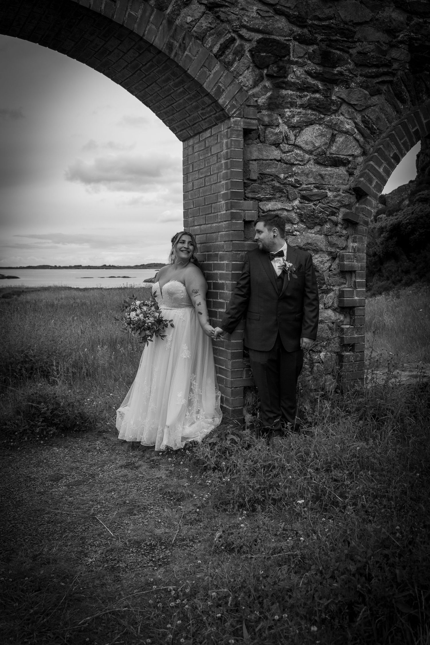Black and white photo of bride and groom standing under a rustic brick archway outdoors.