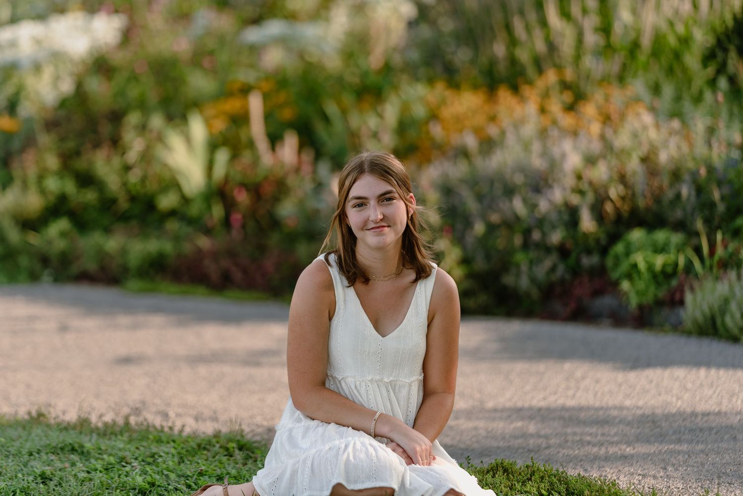 Person in white dress sits on rustic bench with garden flowers in background.